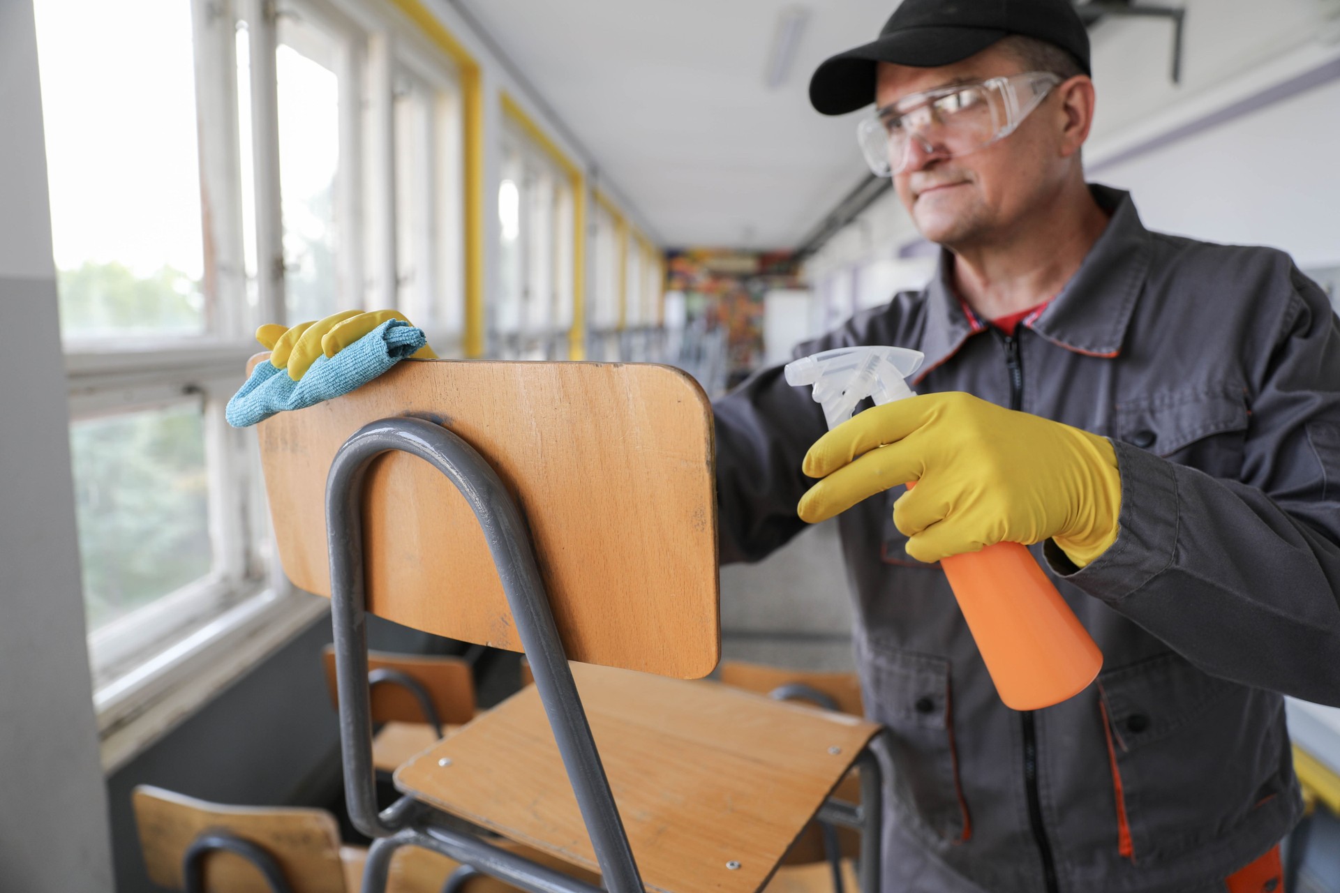 Older Worker Using Handheld Pump Sprayer to Clean Chairs in School